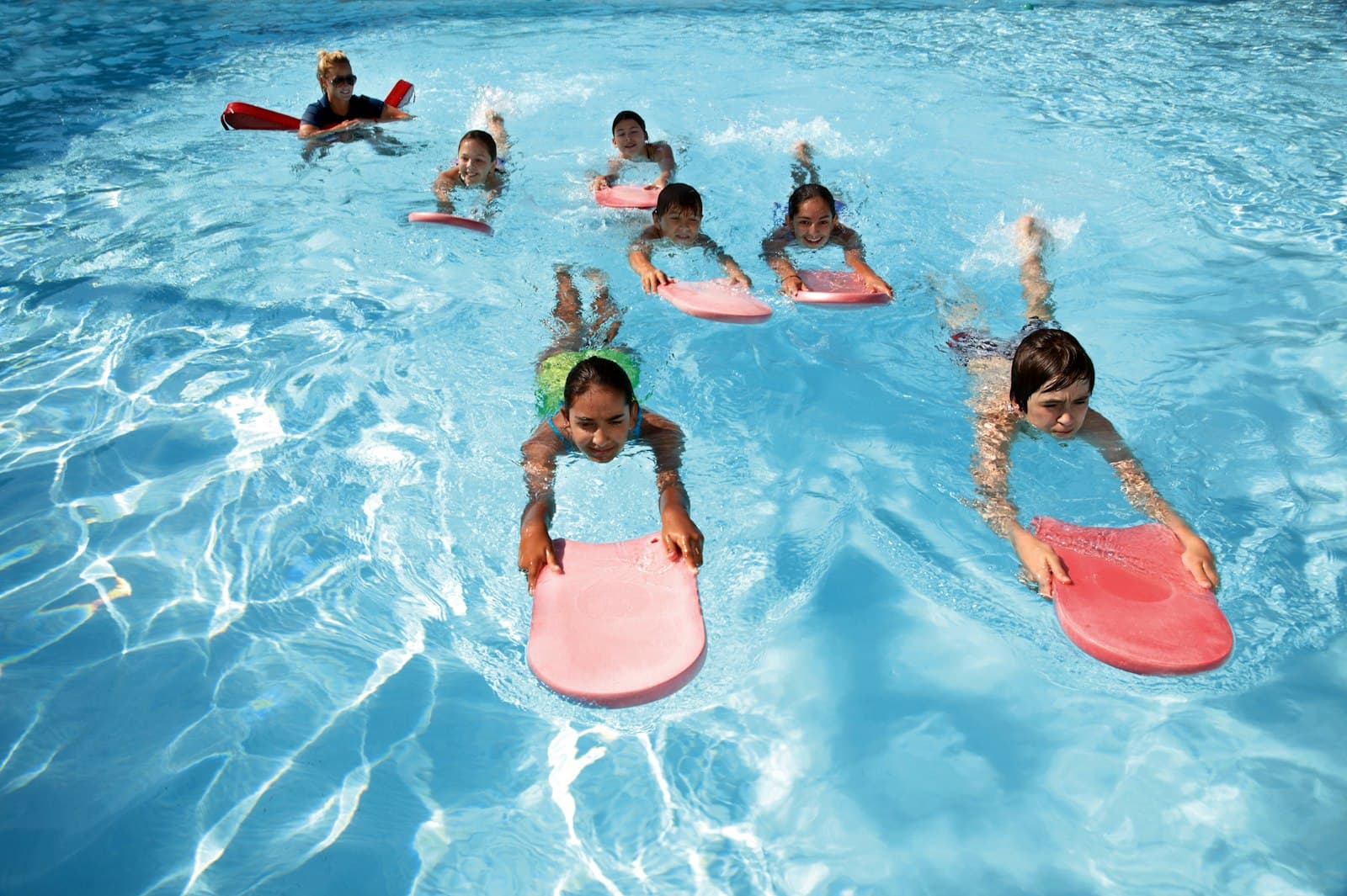 Children on kickboards during a group swim lesson, instructor watching from the back